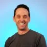Smiling man with short dark hair wearing a dark gray shirt against a blue background.