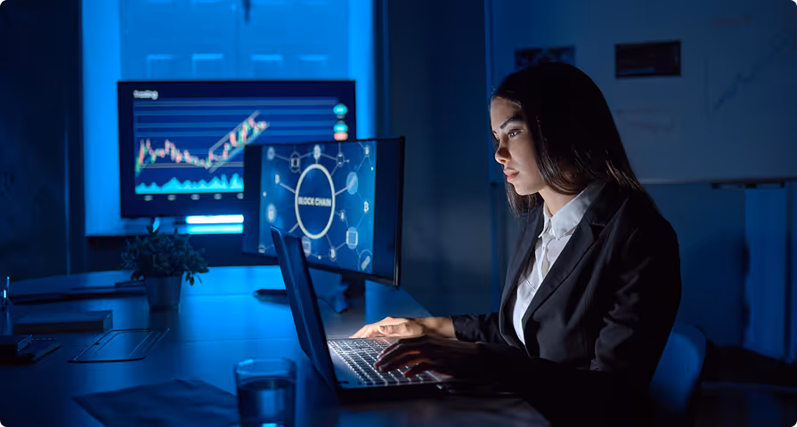 Woman in a dark room working on a laptop with multiple monitors displaying blockchain and trading charts in the background.