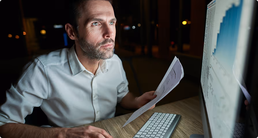Man in a white shirt analyzing documents while looking at a computer screen displaying charts in a dimly lit room.