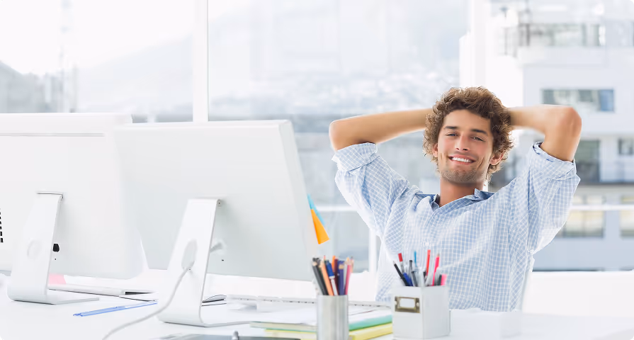 Young man in a blue checkered shirt leaning back with hands behind head smiling at his office desk with computers and stationery.